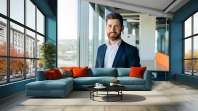 Successful person. Handsome businessman in formal wear standing in office near window, looking and smiling at camera Wall mural