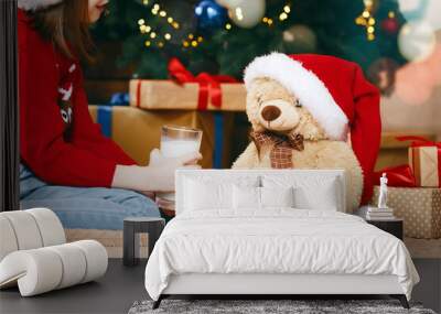 Sweet preschool girl in santa hat feeding teddy bear with cookies and milk next to xmas tree Wall mural