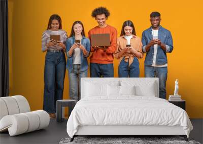 This image shows a group of five young adults standing in a row against a yellow background. They are all looking down at their devices, which include a tablet, a smartphone, and a laptop Wall mural