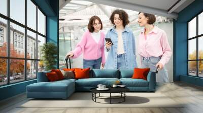 Three travelers in casual attire stand in an airport, smiling while looking at a smartphone. They hold luggage and seem excited about their journey ahead. Wall mural