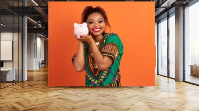 Thrilled pretty black woman holding piggy bank, orange background Wall mural