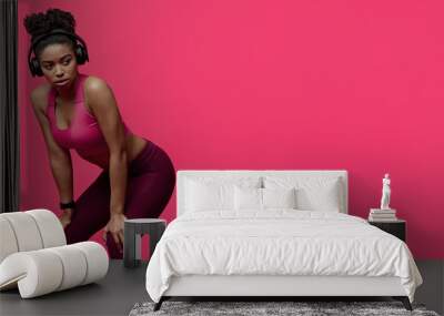 Tired fitness afro girl with wireless earphones having rest, looking at free space, red background, advertising for gym, fitness club Wall mural