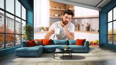 Tired sad sleepy handsome millennial caucasian guy with beard in domestic clothes with cup of coffee sleeps at table Wall mural