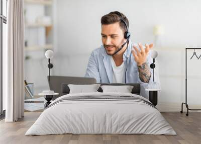 Web conference. Young freelancer man in headset making video call on laptop, sitting at workplace at home Wall mural