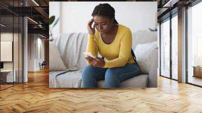 Worried black lady looking at smartphone screen, sitting on couch at home Wall mural