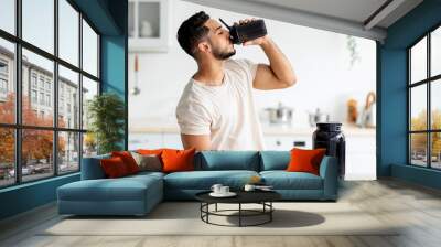 Young Arab guy drinking protein shake from bottle at kitchen, copy space. Millennial Eastern man using meal replacement for weight loss, having sports supplement for muscle gain. Body care concept Wall mural