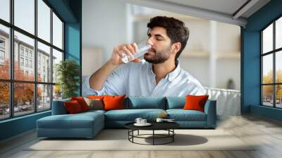 Young arab man drinking fresh water from glass, sitting on sofa in living room interior, copy space Wall mural