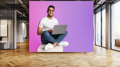 Young Arab man in casual wear working online, sitting cross legged and using laptop in neon light, full length Wall mural