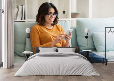 Young Arab Woman Wearing Eyeglasses Relaxing With Smartphone At Home Wall mural