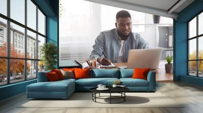 Young businessman using laptop and taking notes at worklace in office Wall mural