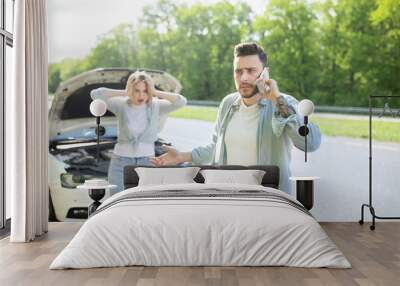 Young guy standing near broken car together with terrified girlfriend, having auto accident, calling breakdown service Wall mural