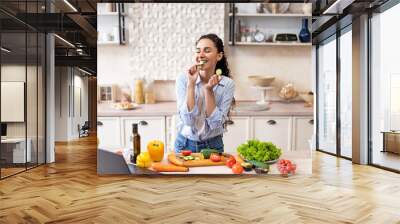 Young latin woman cooking fresh vegetable salad and biting cucumber, preparing lunch at home in kitchen Wall mural