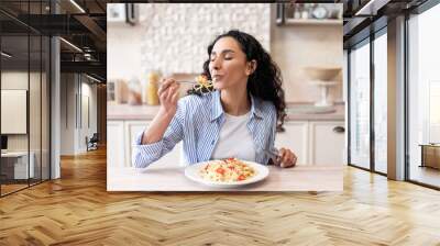 Young latin woman eating delicious pasta, enjoying tasty homemade lunch with closed eyes, sitting in kitchen Wall mural