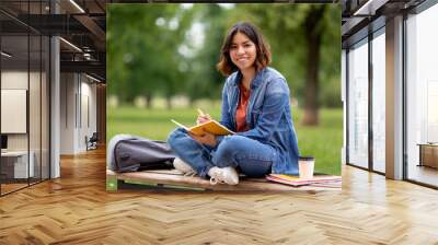 Young middle eastern woman writing in notebook while sitting on bench outdoors Wall mural