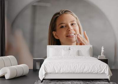 Young woman caring for facial skin using cotton pad and looking in round mirror in bathroom interior Wall mural