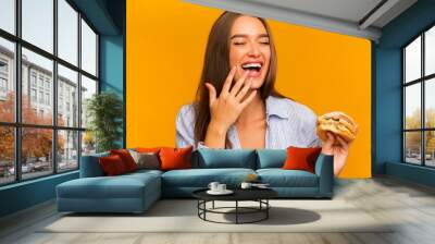 Young Woman Eating Burger And Laughing Standing Over Yellow Background Wall mural