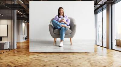 Young woman sitting in armchair, looking at mobile phone screen, using new cool application against white wall Wall mural