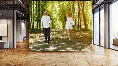 A beautiful and gentle girl with light hair and a white dress is walking in a sunny summer forest with her handsome guy in a white shirt and dark pants and they playing with cute gray dog Wall mural