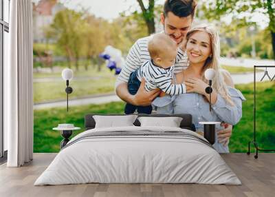 A young and beautiful blonde mother in a blue dress, along with her handsome man dressed in a white jacket, playing with her little son in the summer solar park Wall mural