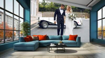 Beautiful bride in a long white dress. Handsome groom in a black suit. Couple near old car Wall mural