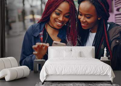 two beautiful and stylish dark-skinned girls with long hair standing in a autumn city near purple wall and using the phone Wall mural