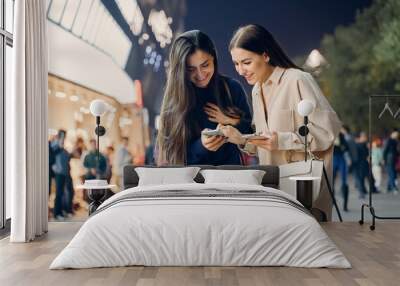 two girlfriends using their cellphone while exploring a new city at night. Two friends searching for information on their cellphone as they are exloring Beijing at night. Girlfriends pointing at an Wall mural