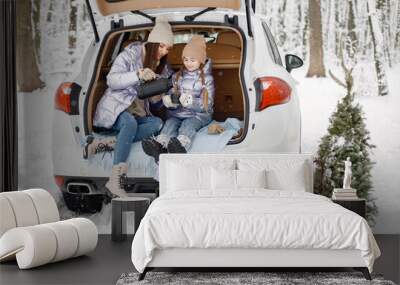 Woman and her daughter sitting in open car's trunk in winter forest Wall mural