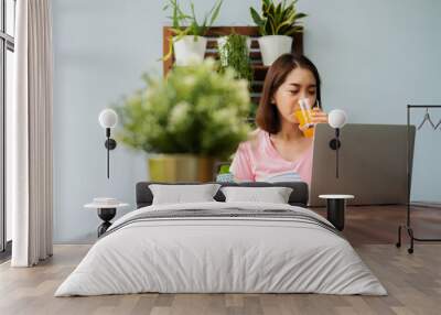 Mid-age Asian woman reading a book and hold orange juice glass in the home. Concept of  Health care And eating for healthy Wall mural