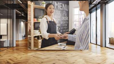 businesswoman customer at coffee shop pays smiling waitress with debit card. young girl barista using tablet while client doing payment on recharge card in cafe counter. happy small business owner. Wall mural