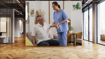 Caring female Asian personal nursing aide helping senior stroke patient do rehabilitation training in the living room at home. she lifts his arm slowly Wall mural