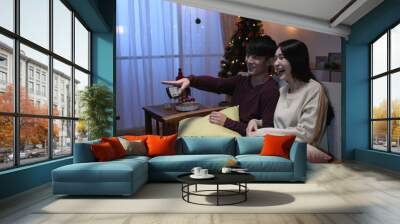 Christmas eve. Loving couple watch tv holding remote control in hand on sofa at home in new year holidays. side view young husband chatting with his wife discussing about talk show and laughing Wall mural