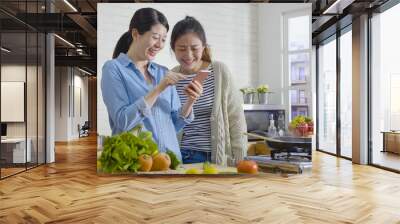 female friends chef cooking together concept. group of asian women roommates making breakfast while checking mobile phone and laughing cheerful. healthy lifestyle friendship prepare meal home kitchen Wall mural