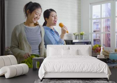 Two cheerful friends cook together and singing with carrot as microphone. asian girl sisters having fun dancing in kitchen with fresh vegetables. happy women laughing joyfully while prepare meal. Wall mural