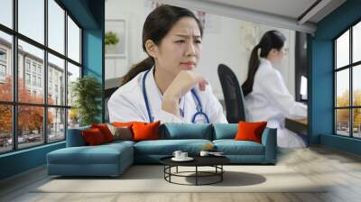 Young asian woman doctor sitting at working desk looking computer screen with serious face thinking about question. female medical staff confused of idea staring at monitor. girl put hand on chin. Wall mural