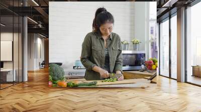 young single lady in modern home kitchen cooking food for dinner. asian chinese woman cutting vegetables by knife on wooden board. girl standing in city apartment chopping cucumber for healthy meal. Wall mural