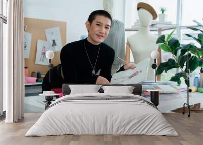 Asian LGBTQ gay dressmaker sketching new fashion dress design on sketch book in studio, standing behind working desk, looking and smiling at camera, small tailor business entrepreneur concept Wall mural