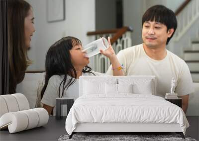 Happy Asian family, father and mother looking at little 6 year old daughter drinking fresh milk from glass, sitting on sofa at home. Selective focus on kid Wall mural