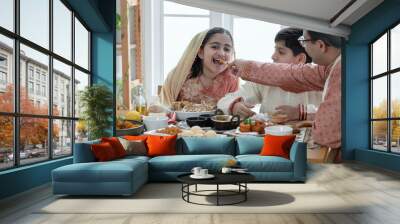 Happy Indian family enjoy eating food with hands, selective focus on Asian cute girl eating naan bread dipping in curry from father feeding, wear traditional clothes, sitting with brother at home Wall mural