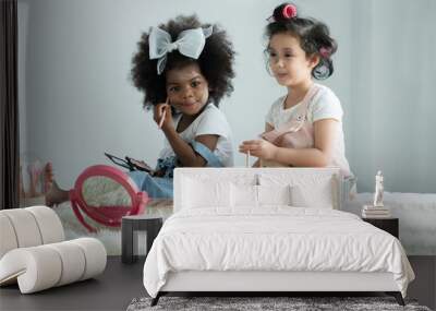 Two little girls friends, African and Caucasian, playing cosmetics together and use pink makeup brushes paint faces, lips, eyes, and cheeks with fun on bed at home. Ethnic diversity kids relationship Wall mural