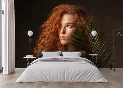 Energizer beautiful young curly ginger hair woman with green tropical plant palm branch posed in dark walls studio. Copy space background. Wall mural