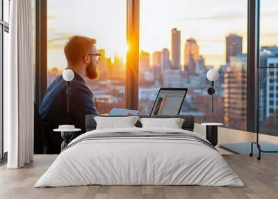 A man sits at a desk, working on his laptop, while the sun sets over the city skyline through a large window. Wall mural