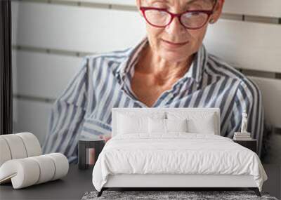 Mature woman with mobile phone on her hands sitting in room and sending messages to her friends and family Wall mural