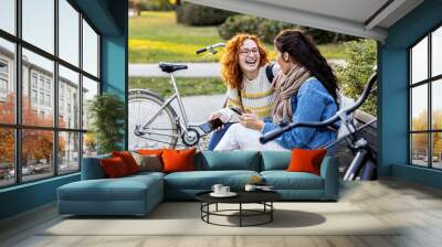 Two girl friends sitting on bench, looking at mobile phone and smile in public par with bycicle on background Wall mural