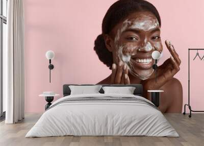 Close up portrait of beautiful black girl wash her face with cleansing face foam. Happy young woman looking at camera. Concept of face skin care. Isolated on pink background. Studio shoot Wall mural