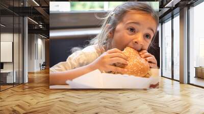 A little girl eats an appetizing burger, close-up. Wall mural