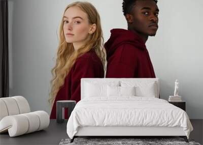 A young woman with long blonde hair and a young man with short black hair stand back-to-back wearing matching burgundy hoodies. They are looking at the camera. Wall mural