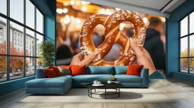 Close-up of a soft pretzel covered in salt, held by two hands in front of a blurred festive background. Wall mural