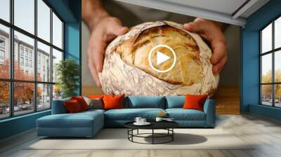 Closeup of a freshly baked sourdough loaf held by hands, with a play button overlayed in the center. Wall mural