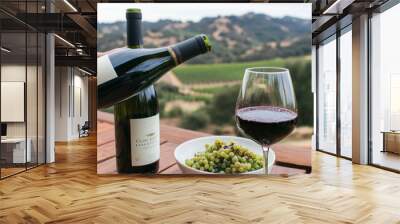 Two bottles of wine, a glass of red wine, and a bowl of green grapes on a wooden table with a vineyard and rolling hills in the background. Wall mural