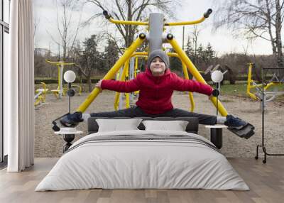 Active small boy 7 years old wearing a red jacket doing sport exercises on a sport athletic playground outdoor in city park at late autumn. Cute boy in split outdoor like Van Damme. Concept of sport. Wall mural